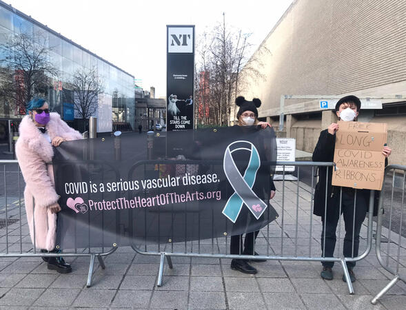 Protect the Heart of the Arts banner Three members of Protect the Heart of the Arts wearing respirators, holding up a banner that reads "Covid is a serious vascular disease" with the Long Covid awareness ribbon and link/QR code to protecttheheartofthearts.org