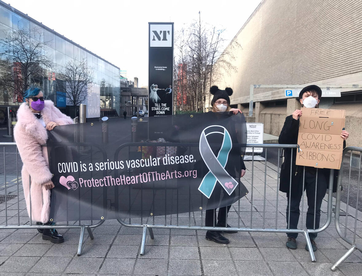 Three members of Protect the Heart of the Arts wearing respirators, holding up a banner that reads "Covid is a serious vascular disease" with the Long Covid awareness ribbon and link/QR code to protecttheheartofthearts.org